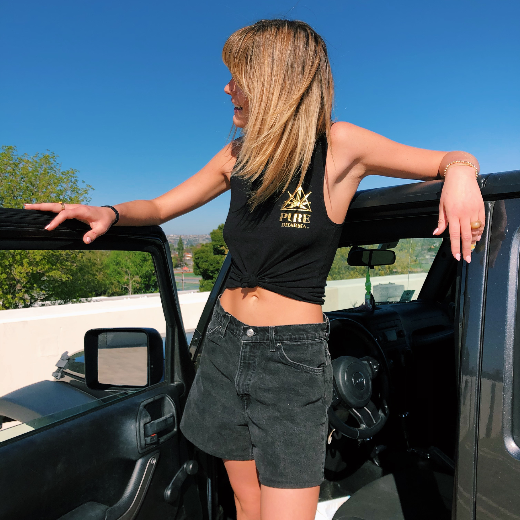 Blond Haired Girl Standing on the Car Door Wearing a Black Pure Dharma Tank Tied Up to be Cropped